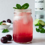 A glass of cherry mojito topped with fresh mint, in the background there are fresh cherries, a bottle of rum, and limes.