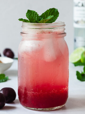 A glass of cherry mojito topped with fresh mint, in the background there are fresh cherries, a bottle of rum, and limes.