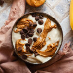 Banana nice cream in a bowl topped with swirls of peanut butter and chocolate chips next to a pink linen napkin.