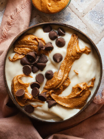 Banana nice cream in a bowl topped with swirls of peanut butter and chocolate chips next to a pink linen napkin.