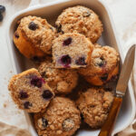 Blueberry muffins stacked decoratively in a baking dish with one split in half next to a knife.