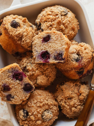 Blueberry muffins stacked decoratively in a baking dish with one split in half next to a knife.
