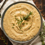 A bowl of mashed butter beans garnished with fresh rosemary and thyme on a white napkin.
