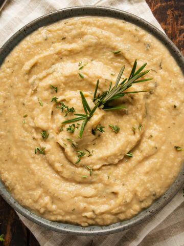 A bowl of mashed butter beans garnished with fresh rosemary and thyme on a white napkin.