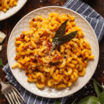 A plate of pan fried butternut squash pasta topped with parmesan cheese and fried sage leaves, on a blue and white striped napkin next to a fork and a small bowl of red chili flakes.