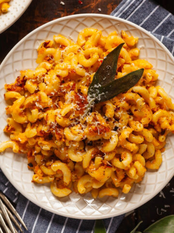 A plate of pan fried butternut squash pasta topped with parmesan cheese and fried sage leaves, on a blue and white striped napkin next to a fork and a small bowl of red chili flakes.