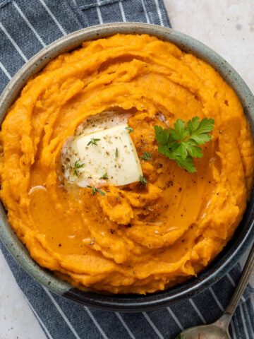 Whipped sweet potatoes garnished with butter, thyme, and parsley in a bowl on top of a blue and white striped napkin.