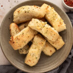 A pile of garlic butter breadsticks on a plate next to a small dish of marinara sauce for dipping.