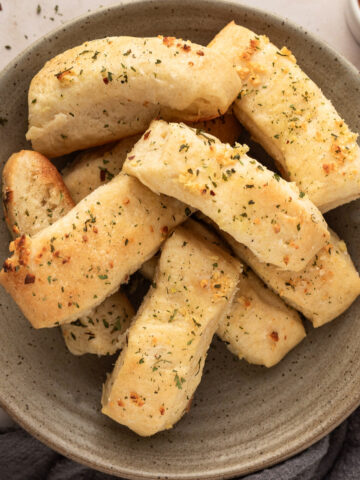 A pile of garlic butter breadsticks on a plate next to a small dish of marinara sauce for dipping.