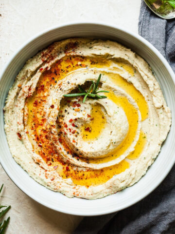 A bowl of white bean dip topped with olive oil, cayenne pepper, and a sprig of rosemary.