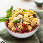 A bowl of sun dried tomato pasta salad on a green linen napkin garnished with fresh basil.