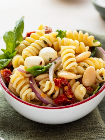 A bowl of sun dried tomato pasta salad on a green linen napkin garnished with fresh basil.