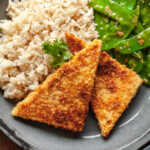 Two slices of crispy pan fried peanut tofu on a plate next to rice and garlic snow peas.
