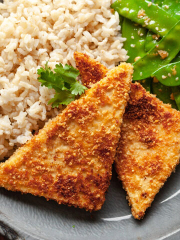 Two slices of crispy pan fried peanut tofu on a plate next to rice and garlic snow peas.