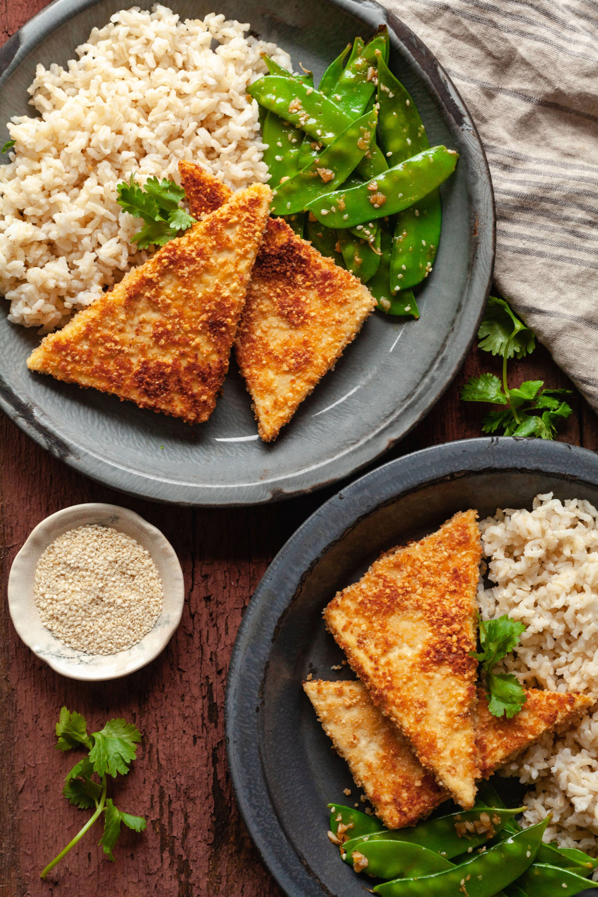 Two plates of two slices of crispy pan fried peanut tofu on a plate next to rice and garlic snow peas.