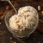 Two scoops of butter pecan ice cream in an ice cream dish with a spoon in it. Ice cream cones and more ice cream dishes are in the background.