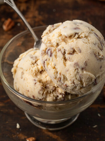Two scoops of butter pecan ice cream in an ice cream dish with a spoon in it. Ice cream cones and more ice cream dishes are in the background.