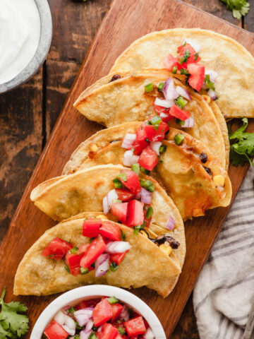 Crispy baked black bean tacos on a serving platter topped with pico next to a bowl of pico and a bowl of sour cream.