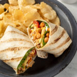 A buffalo chickpea wrap cut in half on a plate next to a pile of potato chips.