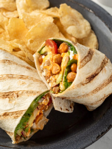 A buffalo chickpea wrap cut in half on a plate next to a pile of potato chips.