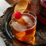 Maple old fashioned garnished with a maraschino cherry on a metal tray next to half an orange and a bottle of bitters.