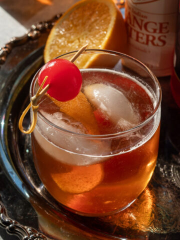 Maple old fashioned garnished with a maraschino cherry on a metal tray next to half an orange and a bottle of bitters.
