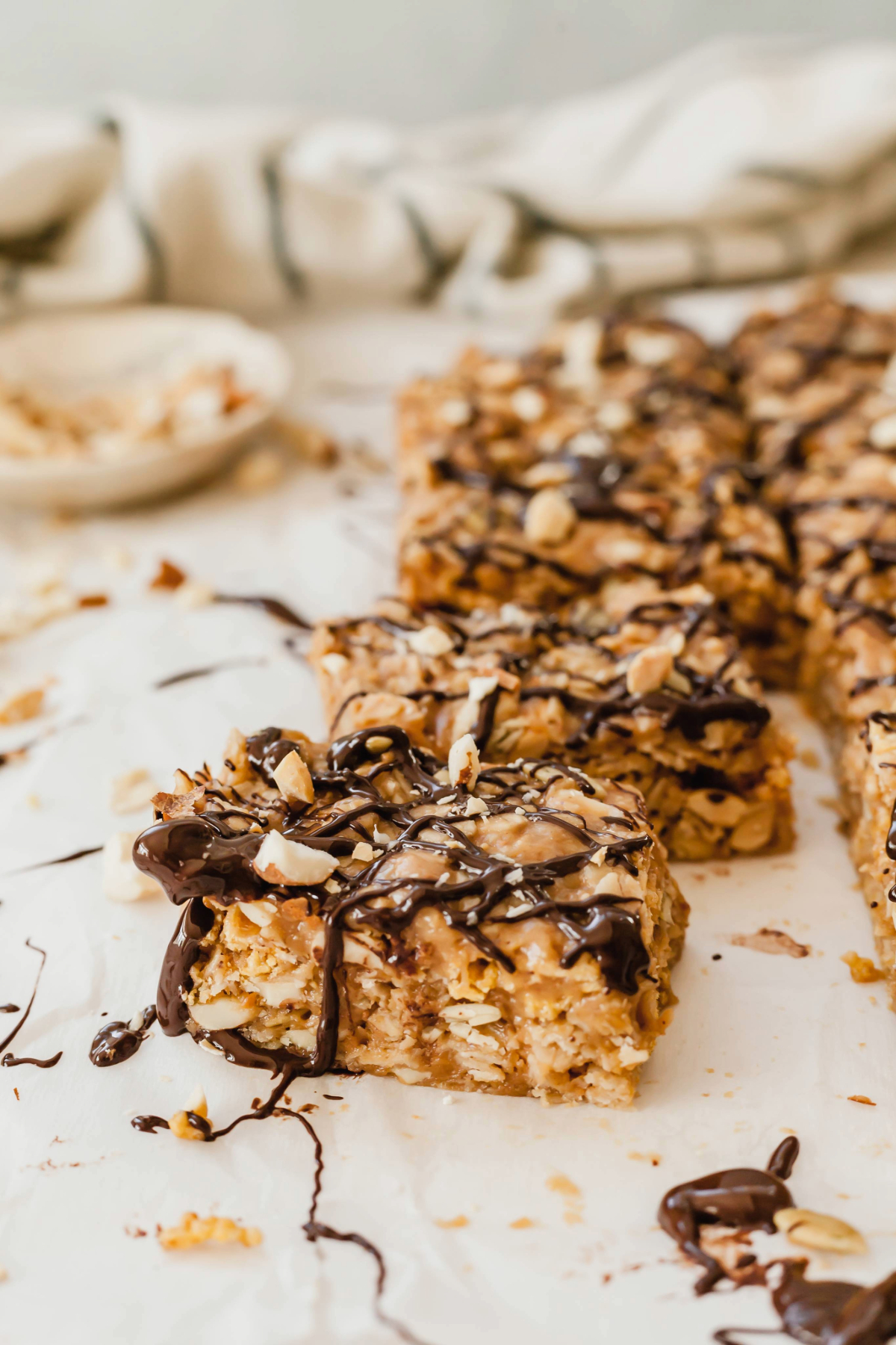 Healthy peanut butter breakfast bars on a sheet of parchment paper next to a bowl of peanuts and a white and black checked cloth napkin.