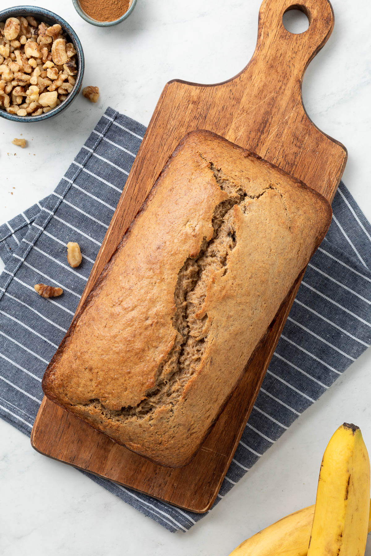 A loaf of cinnamon swirl banana bread on a cutting board, in the background you can see a bunch of bananas and a white and blue striped cloth napkin.