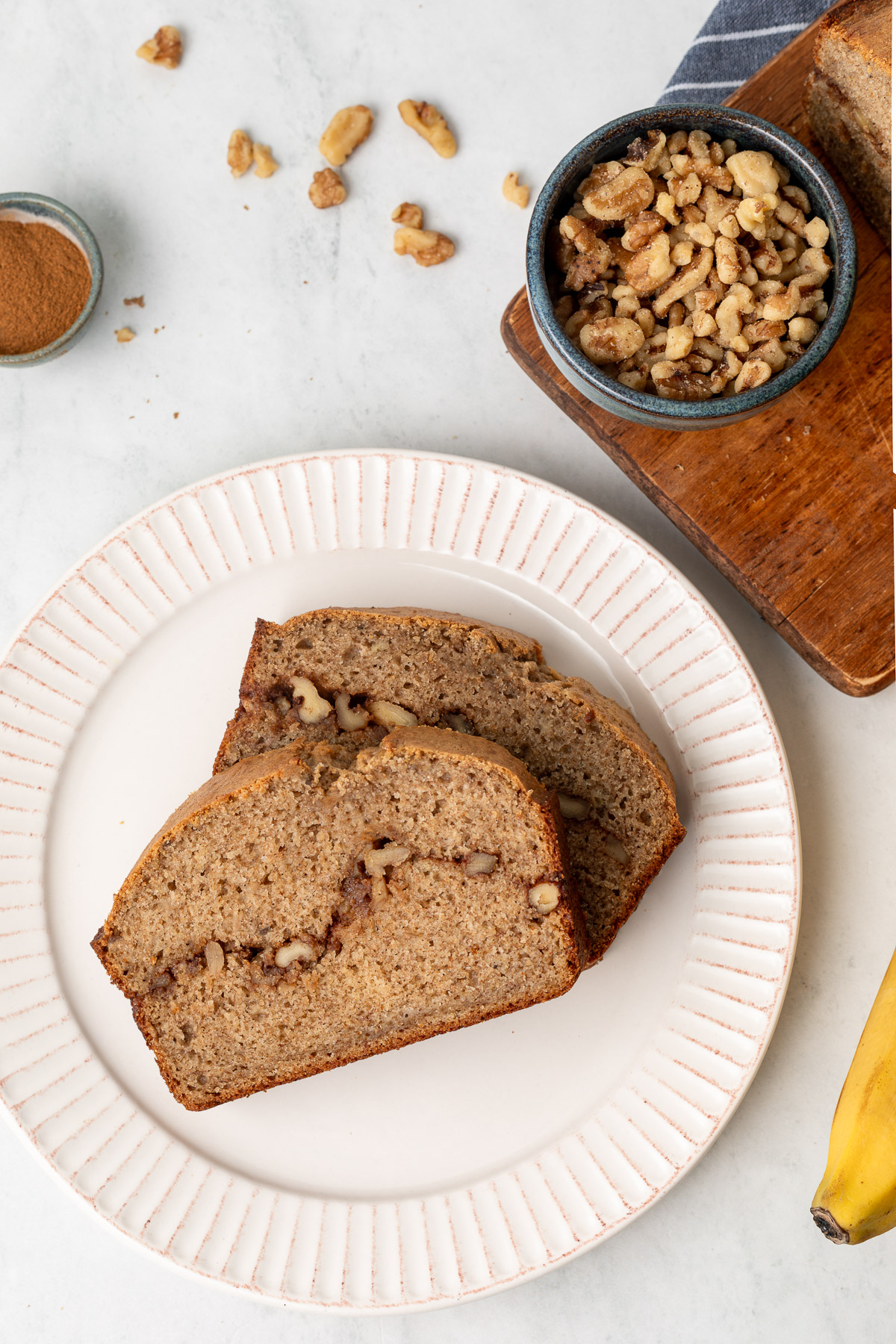 Two slices of cinnamon swirl banana bread on a white plate next to a bowl of walnuts and a cutting board.