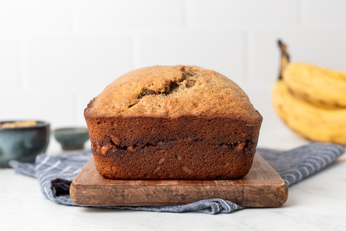 A loaf of cinnamon swirl banana bread on a cutting board, in the background you can see a bunch of bananas and a white and blue striped cloth napkin.