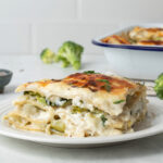 A slice of veggie lasagna on a plate, in the background is the baking dish and broccoli florets.
