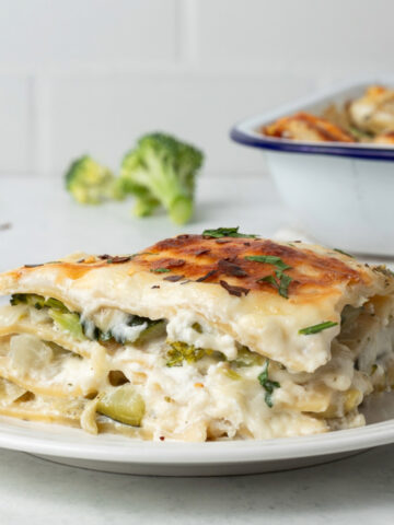 A slice of veggie lasagna on a plate, in the background is the baking dish and broccoli florets.