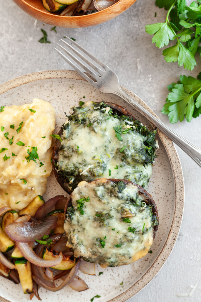 Two spinach stuffed portobello mushrooms on a plate next to a serving of mashed potatoes and sautéed vegetables.