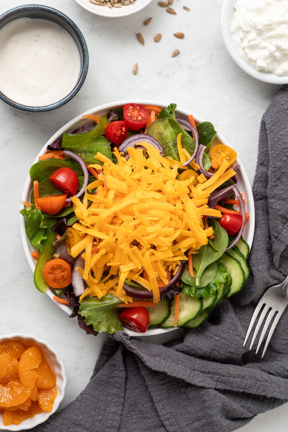 Top down view of a Salad Bar Salad drizzled with no ranch dressing in a white plate next to a gray linen napkin. In the background you can see the ingredients that went into the salad portioned into small bowls.