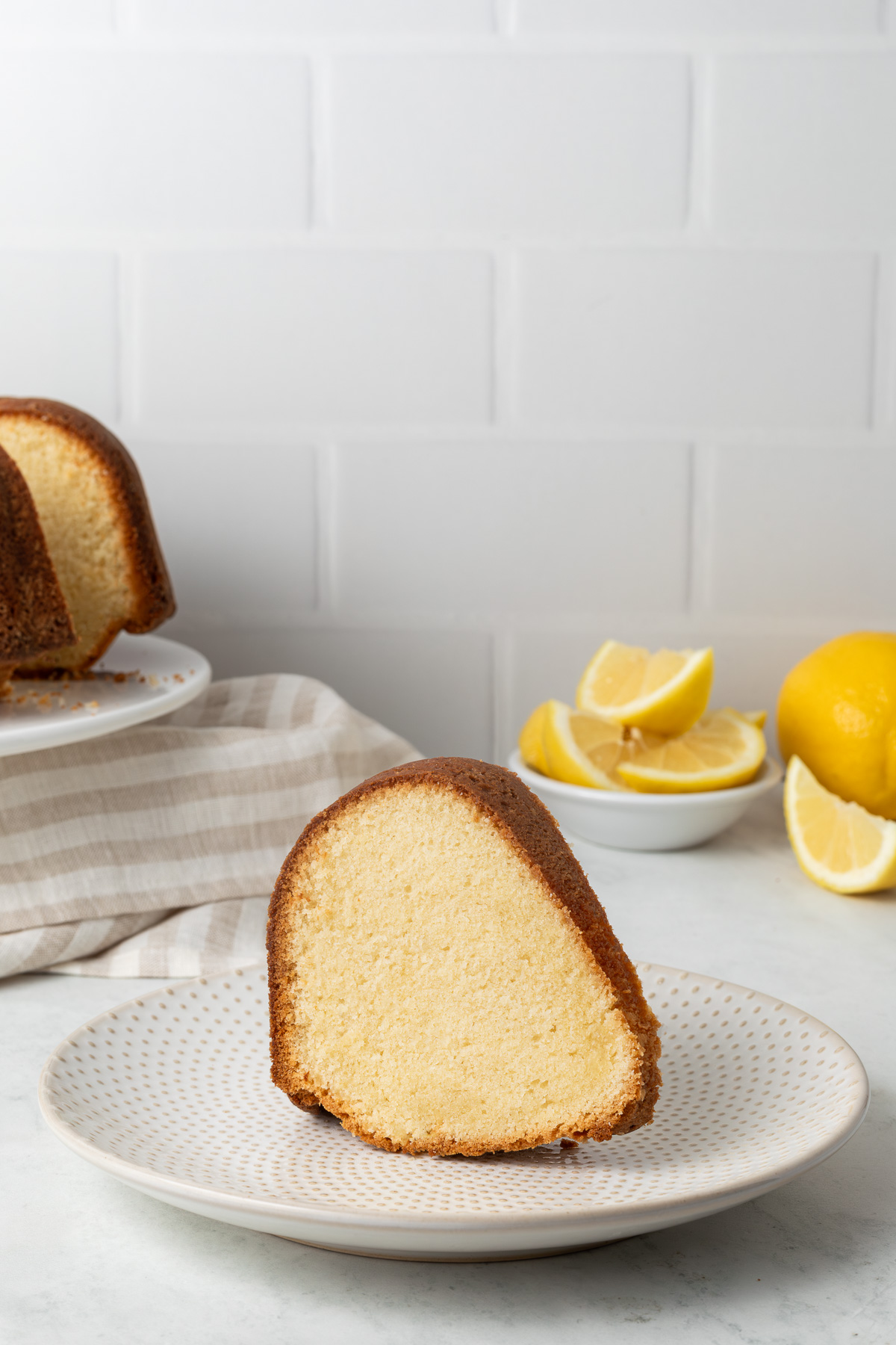 A slice of lemon pound cake next to a cake stand and a bowl of sliced lemons.