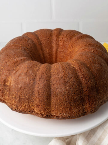 A slice of lemon pound cake next to a cake stand and a bowl of sliced lemons.