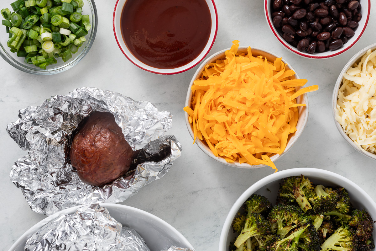 Various toppings in individual bowls next to a baked potato wrapped in foil.
