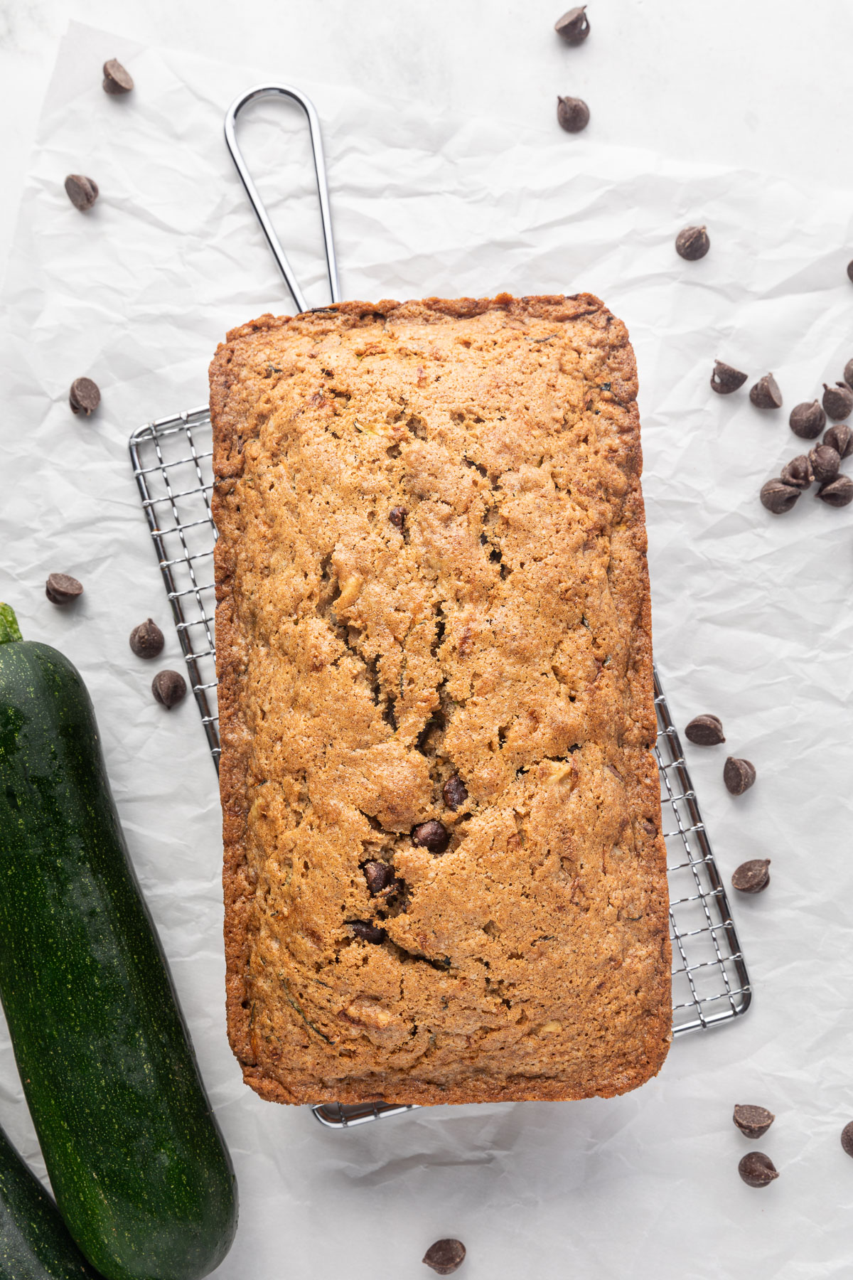 A loaf of chocolate chip zucchini bread on a small cooling rack surrounded by chocolate chips and a zucchini.
