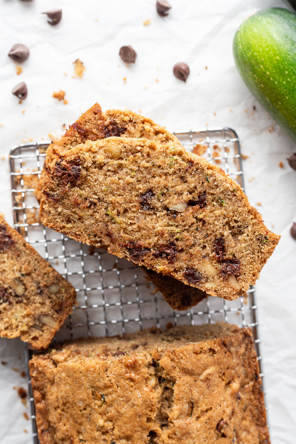Slices cut out of a loaf of chocolate chip zucchini bread on a small cooling rack surrounded by chocolate chips and a zucchini.