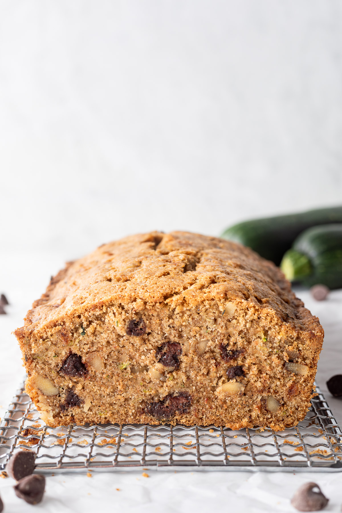 A loaf of chocolate chip zucchini bread on a small cooling rack surrounded by chocolate chips and a zucchini, a slice is cut of the end so you can see inside. 