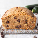 A loaf of chocolate chip zucchini bread on a small cooling rack surrounded by chocolate chips and a zucchini, a slice is cut of the end so you can see inside.
