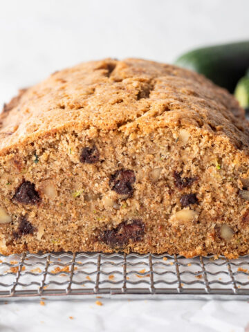 A loaf of chocolate chip zucchini bread on a small cooling rack surrounded by chocolate chips and a zucchini, a slice is cut of the end so you can see inside.