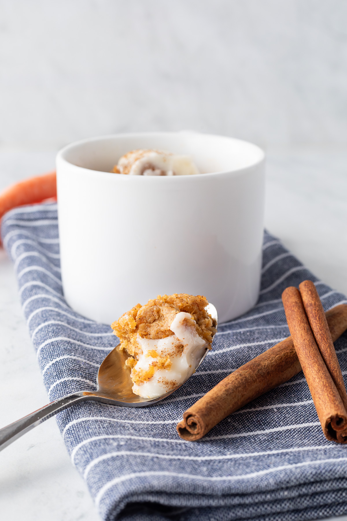 A white mug on a blue and white stripped napkin next to a spoon with carrot cake on it and two cinnamon sticks.