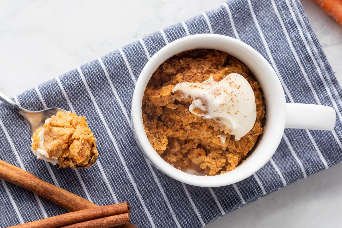 A white mug on a blue and white stripped napkin next to a spoon with carrot cake on it and two cinnamon sticks.