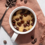 Chocolate chip cookie in a white mug on a pink cloth napkin next to a spoon filled with chocolate chips.