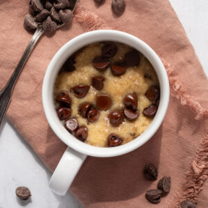 Chocolate chip cookie in a white mug on a pink cloth napkin next to a spoon filled with chocolate chips.