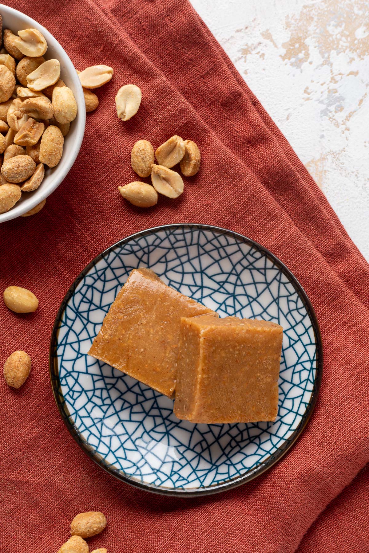 Two squares of peanut butter freezer fudge on a blue pattered plate on a red napkin next to a bowl of peanuts.