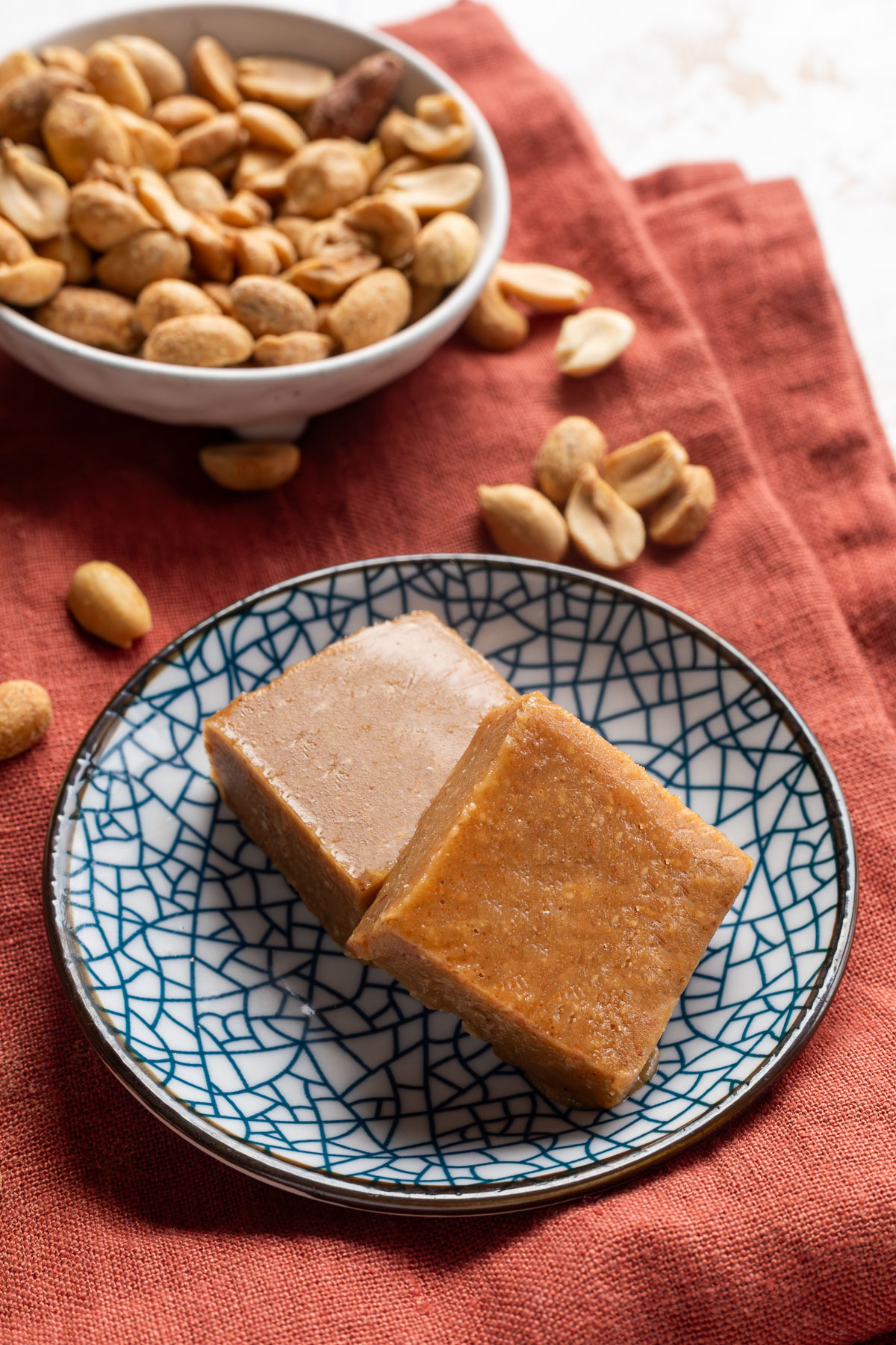 Two squares of peanut butter freezer fudge on a blue pattered plate on a red napkin next to a bowl of peanuts.