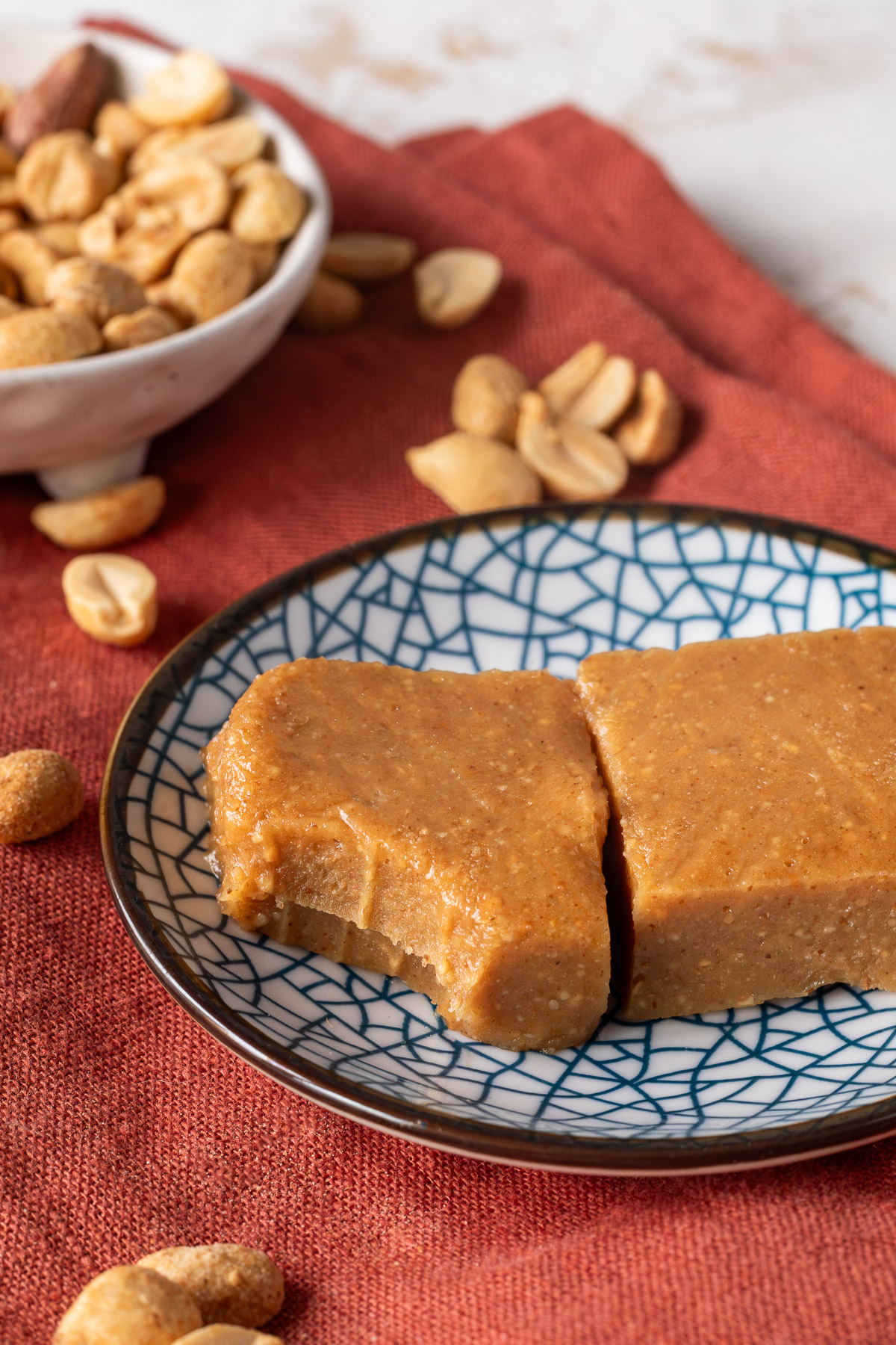 Two squares of peanut butter freezer fudge on a blue pattered plate on a red napkin next to a bowl of peanuts, one of the squares has a bite taken out of it.