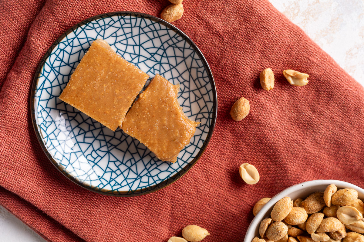 Two squares of peanut butter freezer fudge on a blue pattered plate on a red napkin next to a bowl of peanuts, one of the squares has a bite taken out of it.